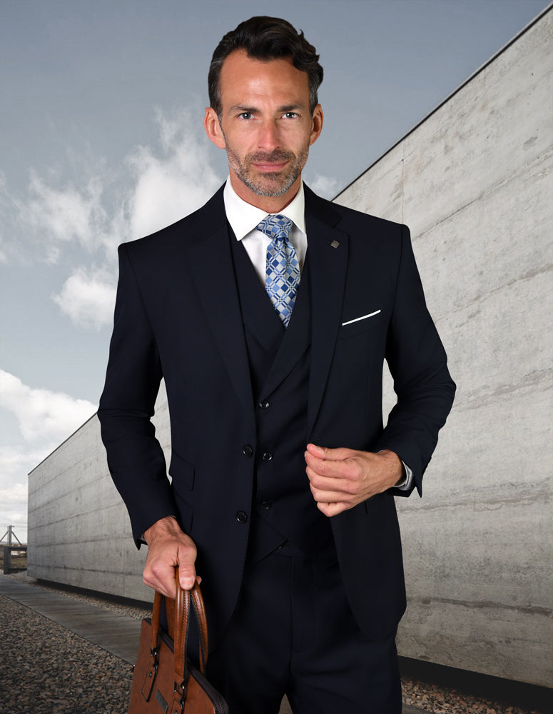 A man wearing the STATEMENT CLOTHING | LAZIO-NAVY by Statement Clothing, featuring tailored flat front pants crafted from luxurious Super 150's fabric, stands outdoors holding a brown briefcase against a backdrop of a concrete wall and cloudy sky.