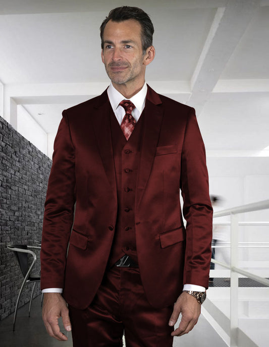 A man in a stylish STATEMENT CLOTHING | LIZA-BURGUNDY slim fit suit from Statement Clothing stands in a modern office setting, with a brick wall and desk visible in the background.