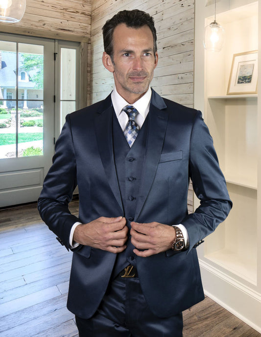 A man in the STATEMENT CLOTHING | LIZA-SAPPHIRE, a slim fit suit from Statement Clothing, stands indoors. He adjusts his jacket made of shiny fabric in a well-lit room with wooden walls and a window in the background.