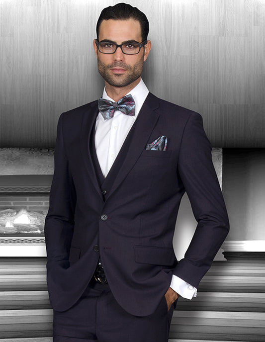 Lorenzo, dressed in the sleek STATEMENT CLOTHING | LORENZO-EGGPLANT suit from Statement Clothing, accessorized with a bow tie and glasses, stands confidently in a modern setting.