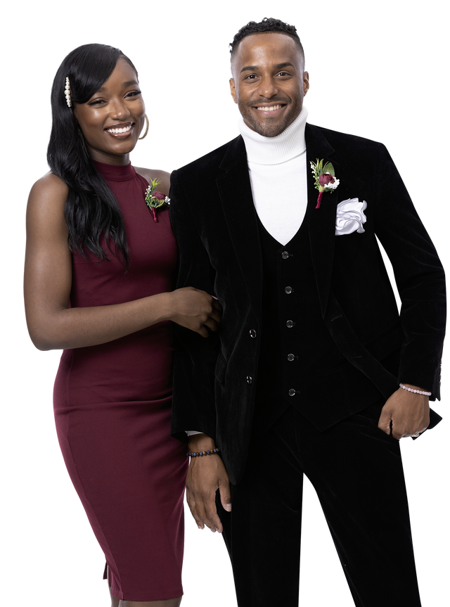 A smiling couple dressed in formal attire showcases fancy fashion, with the woman in a burgundy dress and the man wearing the EJ Samuel Black Suit M2781 paired with a white turtleneck, both adorned with floral boutonnieres.