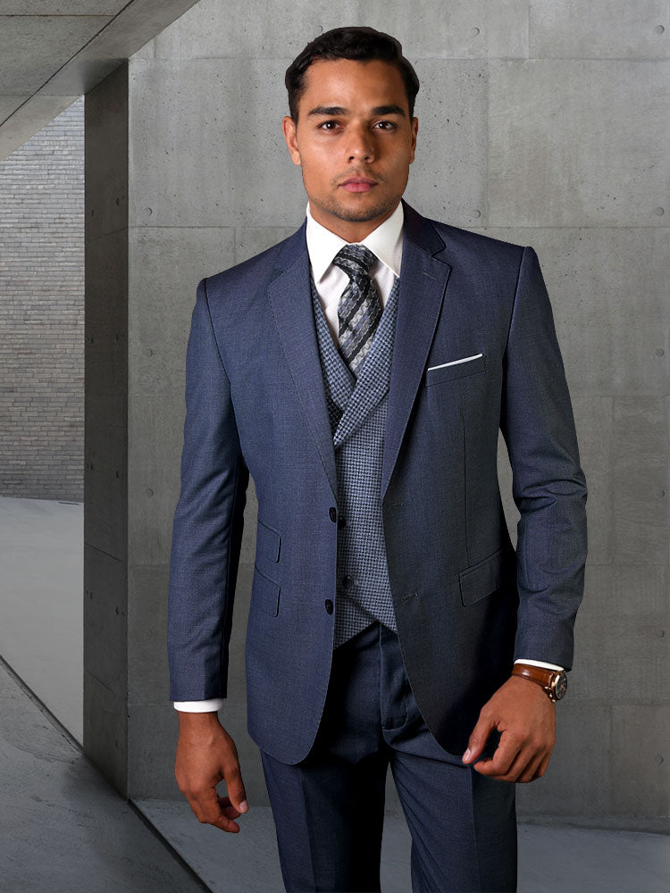 A man in the STATEMENT CLOTHING | MARCO-INDIGO suit crafted from luxurious Italian wool, complemented by a patterned tie, stands confidently against a concrete wall, looking directly at the camera.