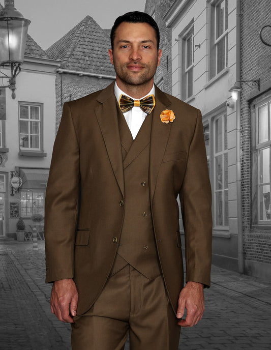 A man dressed in the Messina-Bronze ensemble from Statement Clothing, featuring a brown suit with pleated pants, a double-breasted vest, and accessorized with a bow tie and bronze boutonniere, stands on a cobblestone street against vintage buildings serving as the perfect backdrop.