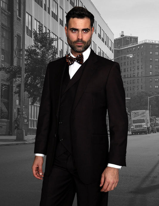 A man in the Statement Clothing's MESSINA-BROWN ensemble, featuring a black suit and bow tie with a double-breasted vest, stands before a city street backdrop.