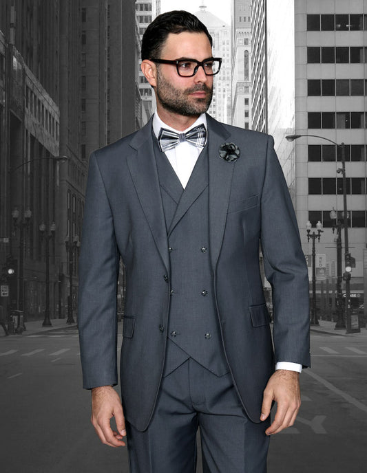 A man dressed in the STATEMENT CLOTHING | MESSINA-CHARCOAL three-piece suit by Statement Clothing, comprising a double-breasted vest and pleated pants, stands on a city street with towering skyscrapers in the background.