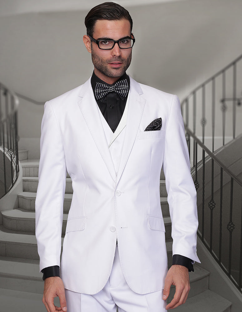 A man adorned in the STATEMENT CLOTHING | MESSINA-WHITE by Statement Clothing is wearing a sharp white suit paired with a black shirt, bow tie, and glasses. He completes his sophisticated look with a double-breasted vest while standing in front of a staircase.