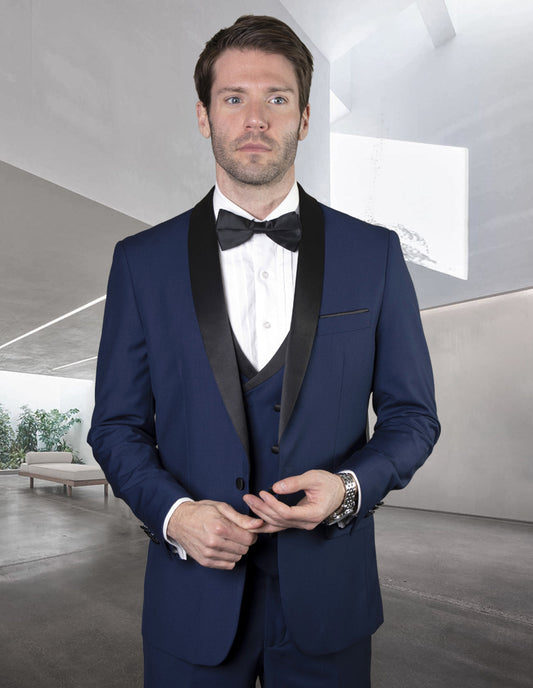 A man wearing the MGM-SAPPHIRE from Statement Clothing, a blue tuxedo suit made of fine Italian fabric and accompanied by a black bow tie, stands indoors against a modern, minimalistic background.