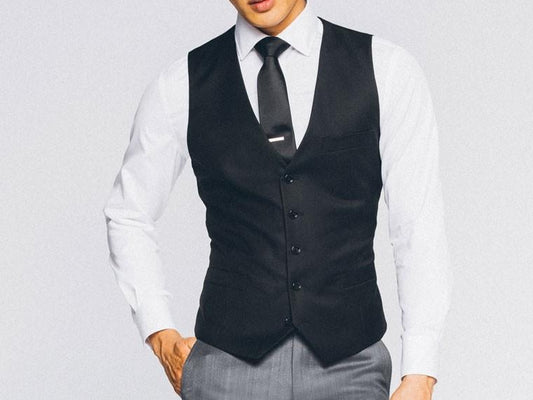 An individual wearing a white shirt, the Vinci Slim Fit Single Breasted 5 Button Vest (Black) OV-900 by Vinci Suits, a black tie, and gray pants stands against a plain background.