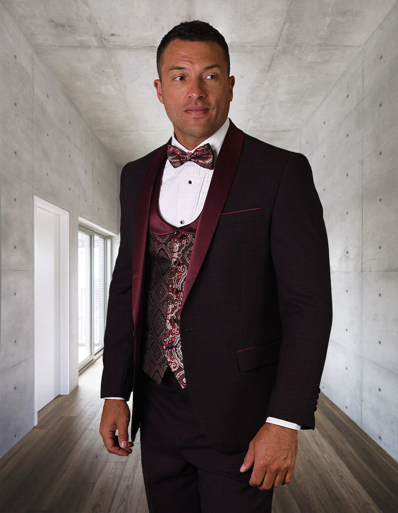 A man wearing the STATEMENT CLOTHING | PALMERO-BURGUNDY tuxedo from Statement Clothing, complete with a patterned waistcoat and bow tie, stands in a modern, concrete-walled hallway.