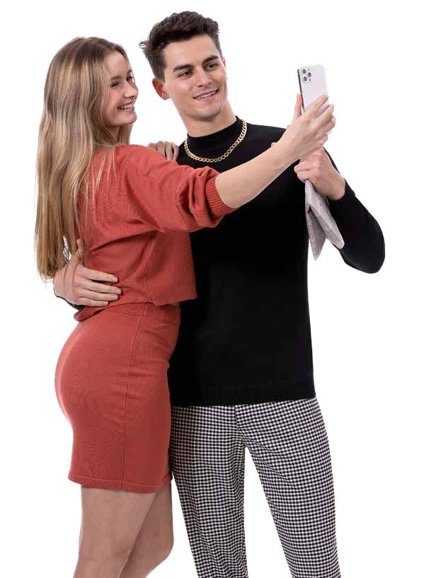 A woman in a red outfit and a man wearing EJ Samuel Blk/White Plaid Pants PL150 with a black sweater take a selfie together, smiling at the phone.