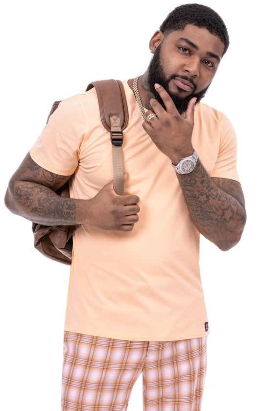 A man with a beard and tattoos, sporting EJ Samuel Pink Plaid Pants PL96 in a modern fit and a peach t-shirt, holds a backpack strap with one hand while thoughtfully touching his chin with the other.