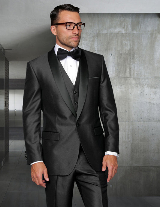 A man wearing a sophisticated slim-fit tuxedo from Statement Clothing stands indoors against a grey wall, accessorized with a shawl lapel and bow tie, while sporting glasses.