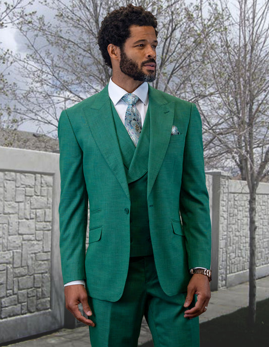 A man in a RIMA-FOREST green wool suit by Statement Clothing, featuring a peak lapel and coordinating double-breasted vest, stands outdoors near a stone wall and leafless trees.