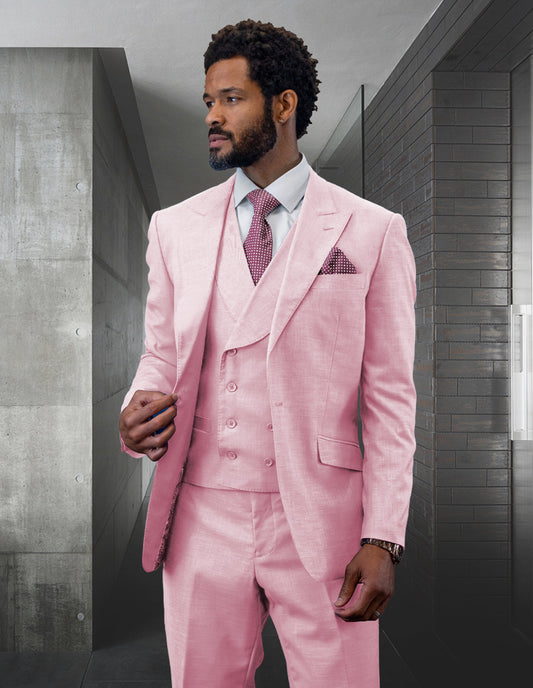 A man wearing the STATEMENT CLOTHING | RIMA-PINK suit from Statement Clothing, paired with a white shirt and pink tie, stands confidently in an industrial-style room with gray walls. The ensemble is highlighted by its stylish peak lapel, adding a touch of elegance to the modern setting.