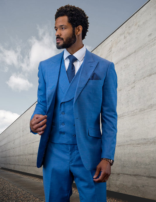 A man wearing the RIMA-SAPPHIRE suit by Statement Clothing with a double-breasted vest and peak lapel stands outdoors against a modern concrete wall, looking to the side.