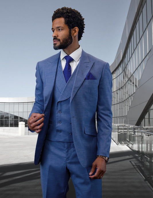 Dressed in the STATEMENT CLOTHING | RIMA-STEEL BLUE from Statement Clothing, a man stands outdoors against a modern glass building. His elegant ensemble, crafted from luxurious Super 180's Wool, includes a sleek blue peak lapel suit with a double-breasted vest and is accented with a polka dot pocket square as he gazes thoughtfully to the side.