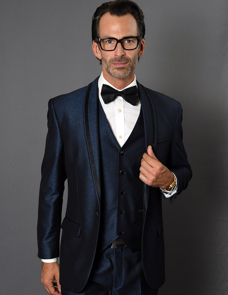 Man in a tailored-fit RIO-INDIGO tuxedo suit from Statement Clothing, complete with a bow tie and glasses, standing against a gray background.