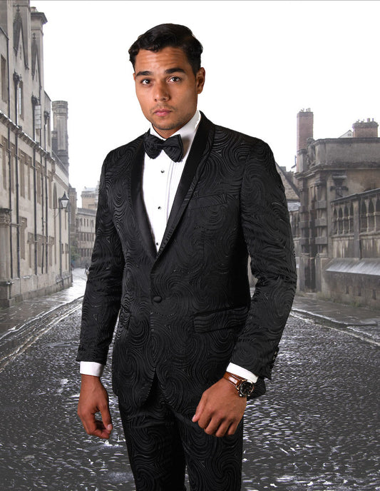 A man in the STATEMENT CLOTHING | RJS-104-BLACK, a slim fit, patterned black tuxedo from Statement Clothing, with a matching bow tie, stands on a cobblestone street with historic buildings in the background.