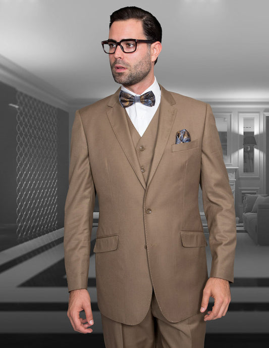 Wearing an elegant tailored fit three-piece suit by Statement Clothing, specifically the STATEMENT CLOTHING | STZV-100-BRONZE crafted from luxurious Super 150's fabric, a man stands confidently with his bow tie and glasses in a modern room featuring geometric decor.