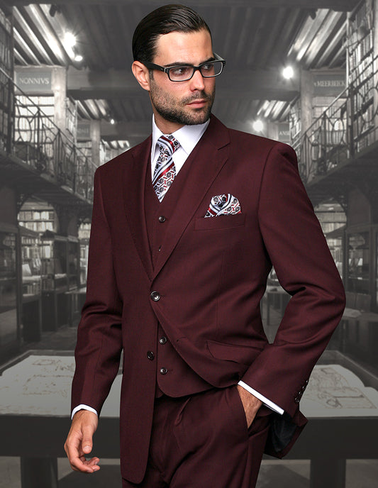 A man in Statement Clothing's STATEMENT CLOTHING | STZV-100-BURGUNDY, a burgundy three-piece suit with tailored fit flat front pants and glasses, stands confidently in a dimly lit library.