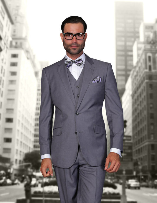 A man wearing the STATEMENT CLOTHING | STZV-100-CHARCOAL suit from Statement Clothing, featuring flat front pants and a tailored fit, stands confidently against the cityscape backdrop of towering buildings. He completes his polished look with glasses and a patterned bow tie.