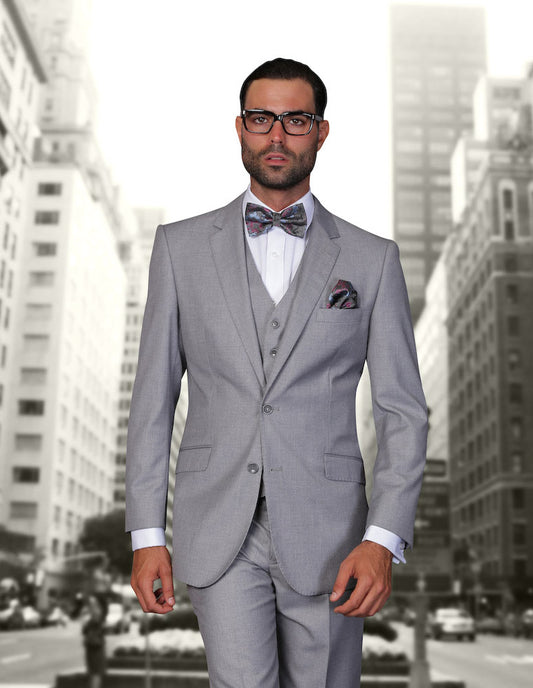 A man confidently stands on a city street lined with tall buildings, wearing the STATEMENT CLOTHING | STZV-100-GREY from Statement Clothing—a stylish gray three-piece suit with flat front pants and a tailored fit, perfectly accessorized with a bow tie and glasses.