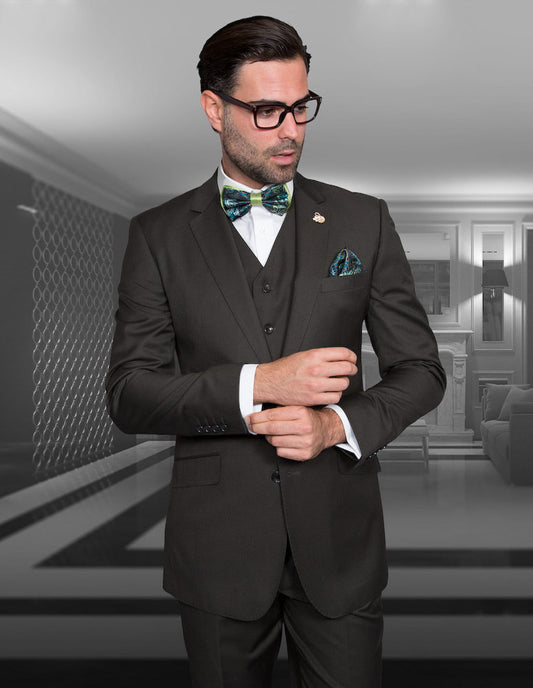 In a contemporary, monochrome room, the man in a dark suit featuring Statement Clothing's STZV-100-OLIVE flat front pants and tailored fit adjusts his sleeve, perfectly complementing his vest and bow tie.