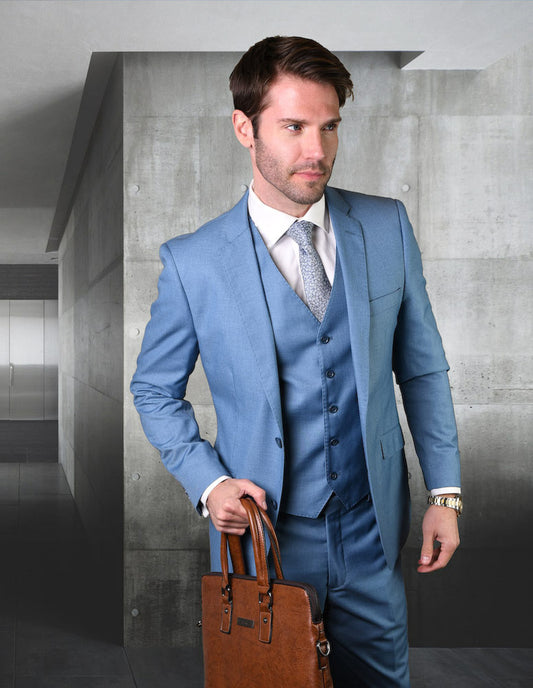 A man dressed in a STATEMENT CLOTHING | STZV-100-STEELBLUE suit from Statement Clothing, featuring flat front pants, holds a brown leather briefcase as he stands in a modern corridor.