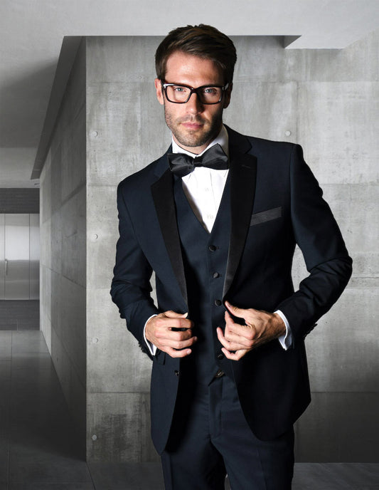 A man in a tailored-fit black tuxedo with glasses and a bow tie stands in a modern hallway with concrete walls, his ensemble perfectly complemented by sleek navy flat front pants from the STATEMENT CLOTHING | T2000PV-NAVY collection by Statement Clothing.