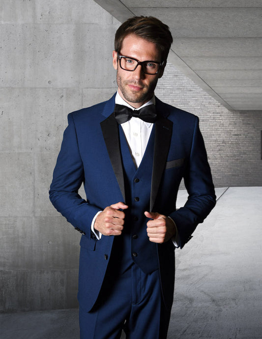 A man in the STATEMENT CLOTHING | T2000PV-SAPPHIRE suit from Statement Clothing, featuring flat front pants and a black bow tie, stands in a concrete hallway holding his jacket. The tailored fit highlights his stature, while the sapphire hue of the fabric adds an extra touch of elegance.