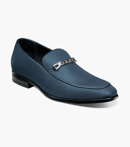 The Stacy Adams TAZIO Moc Toe Bit Slip On in Navy (model 25499-410) showcases a blue textured knit fabric upper adorned with a silver chain detail on the front. This slip-on style includes a memory foam footbed for added comfort, complemented by a black sole and slightly raised heel, all set against a plain white background.