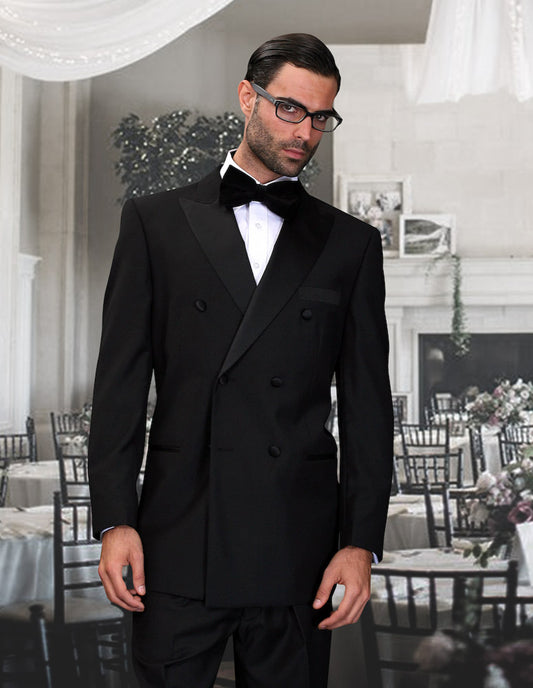 A man dressed in the sophisticated STATEMENT CLOTHING | TUX-DB-BLACK by Statement Clothing is standing in a formal dining setting adorned with beautifully decorated tables and chairs. He accessorizes with glasses and a black bow tie, while his impeccably tailored pleated pants add the ultimate touch of elegance.