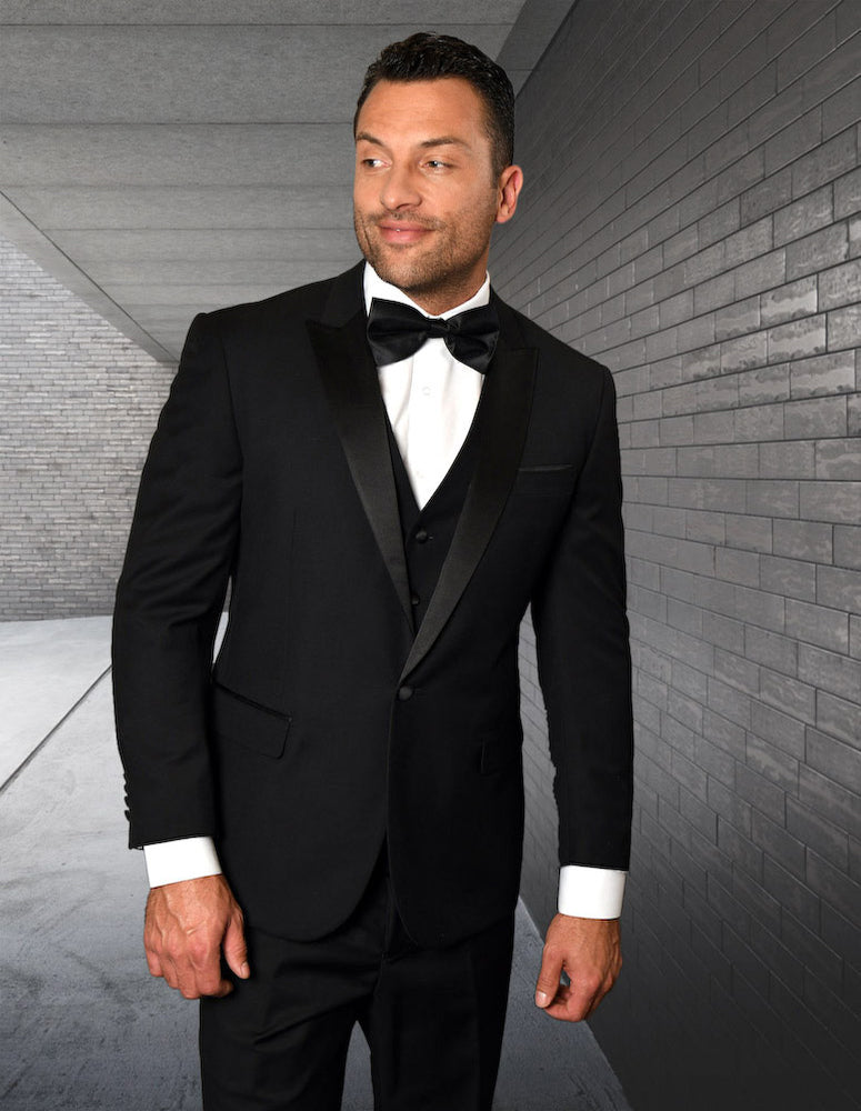 Wearing the STATEMENT CLOTHING | TUX-PK-BLACK from Statement Clothing, a man stands against a gray brick wall in a black peak lapel tuxedo paired with a bow tie, highlighting its sleek, tailored fit and luxurious Italian fabric as he gazes to the side.