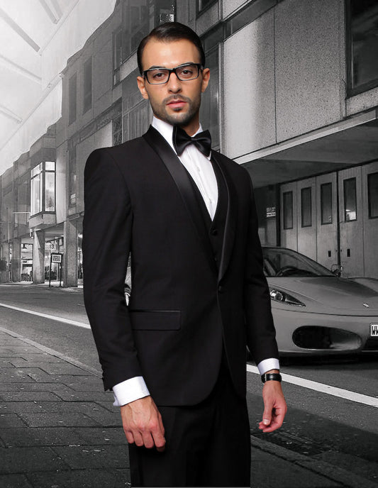 A man in a STATEMENT CLOTHING | TUX-SH-BLACK by Statement Clothing, featuring flat front pants and a tailored fit shawl lapel tuxedo, stands confidently on a city street with a modern building and parked car in the background.