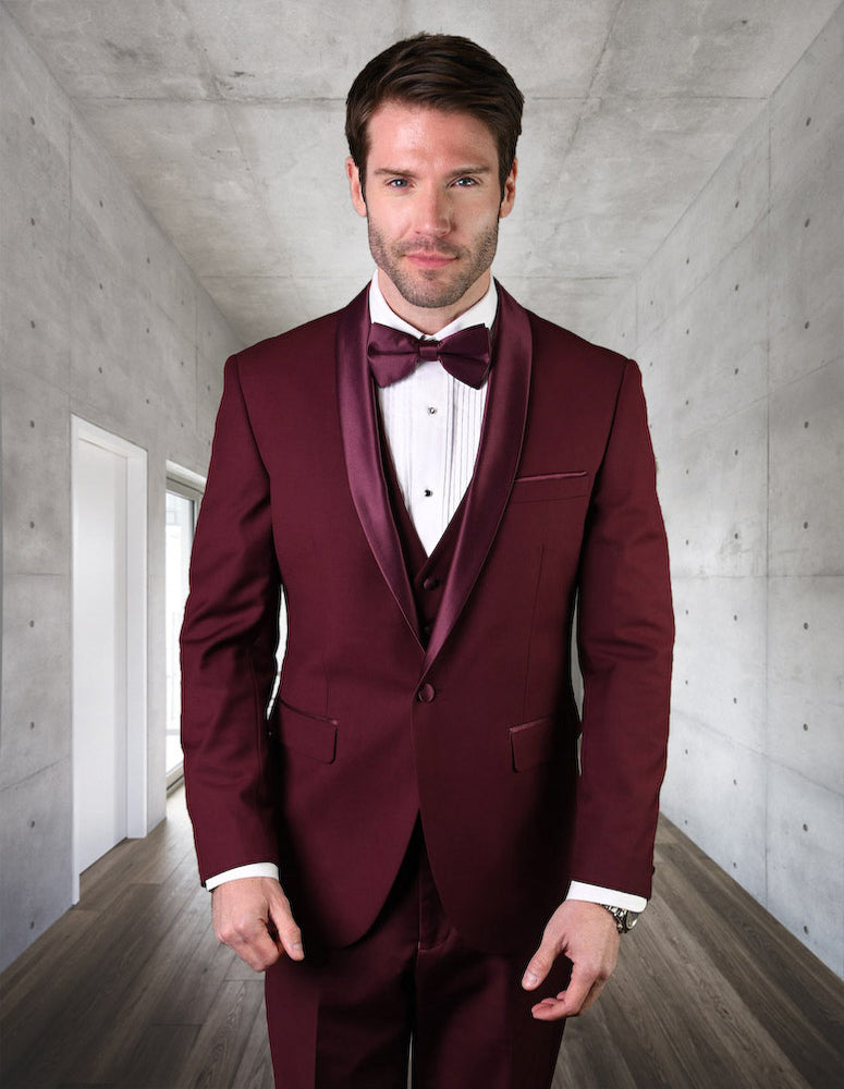 A man in a Statement Clothing tailored fit burgundy tuxedo, product TUX-SH-BURGUNDY, with a matching bow tie stands in a modern hallway with gray walls and wood flooring.