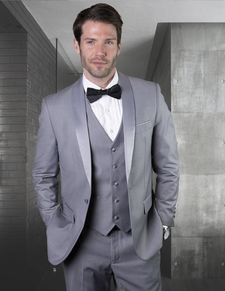 A man wearing the STATEMENT CLOTHING | TUX-SH-GREY-GREY by Statement Clothing, featuring a tailored-fit gray three-piece suit with flat front pants and a bow tie, stands in a modern, minimalist room with concrete and brick walls.