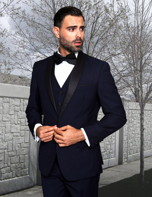 A man in a Statement Clothing TUX-SH-NAVY-BLACK tuxedo and bow tie stands outdoors in front of a wall and bare trees, adjusting his jacket.