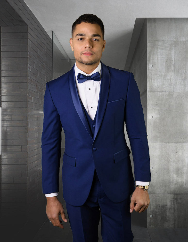 A man in a tailored sapphire blue suit from Statement Clothing's TUX-SH-SAPPHIRE-1 collection, complete with a bow tie, stands in a modern, minimalist hallway, exuding elegance.