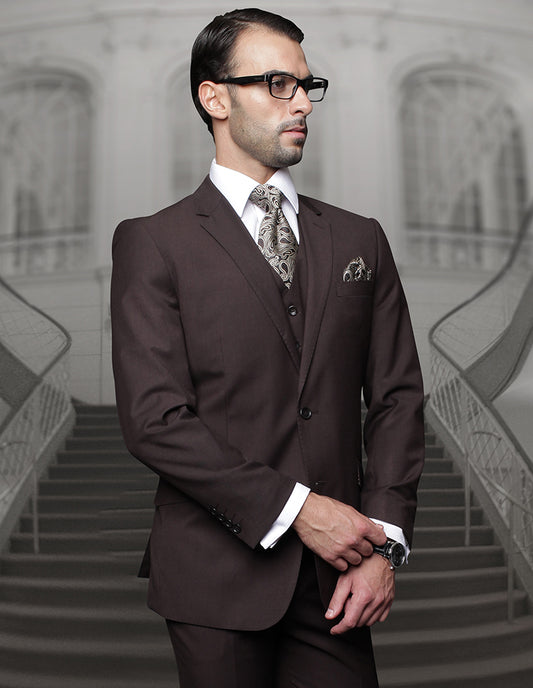 A man in a STATEMENT CLOTHING | TZ-100-BROWN suit from Statement Clothing, paired with pleated pants and glasses, stands confidently in front of a curved staircase, looking to the side.