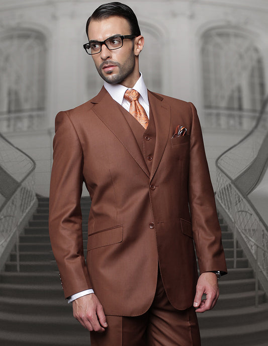 A man wearing the STATEMENT CLOTHING | TZ-100-COPPER from Statement Clothing, featuring a copper-brown three-piece suit and glasses, stands in front of a staircase with an ornate railing, his ensemble completed with Super 150's fabric and regular fit pleated pants.