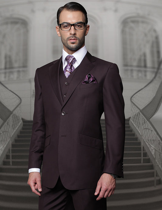 A man in a brown suit by Statement Clothing, featuring regular fit pleated pants, accessorized with a STATEMENT CLOTHING | TZ-100-EGGPLANT tie and pocket square, stands elegantly in front of a staircase backdrop.