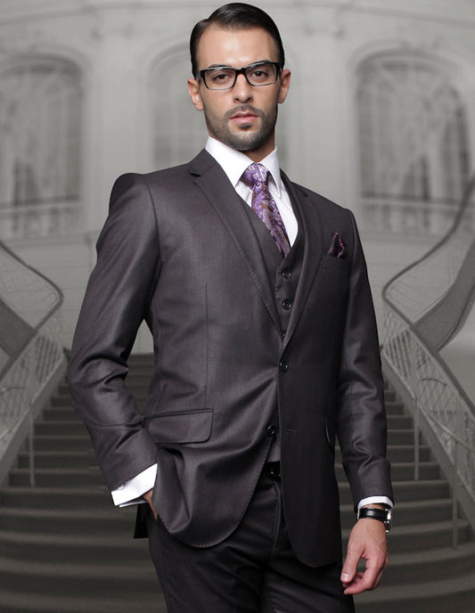 A man in a dark suit and glasses stands confidently, embodying the elegance of Statement Clothing's Super 150's fabric from their STATEMENT CLOTHING | TZ-100-H.CHARCOAL collection. His regular fit trousers with pleated pants add an air of sophistication against the blurred staircase in the background.