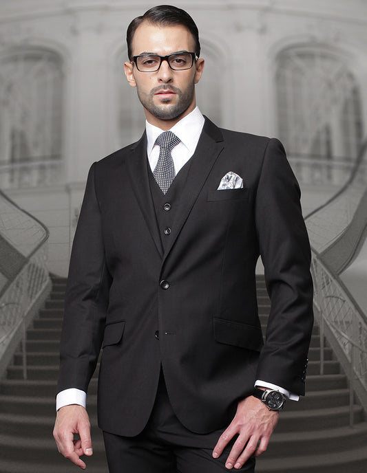 A man confidently stands in front of a staircase, dressed in a black suit from the STATEMENT CLOTHING | TZ-100BG-BLACK by Statement Clothing, made from Super 150s wool. The ensemble includes pleated pants and boasts a regular fit. His neatly knotted tie and glasses add an elegant touch of sophistication.