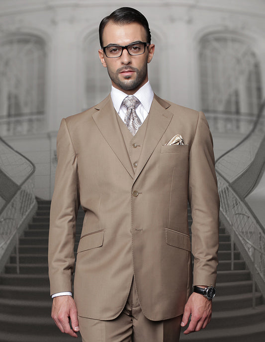 A man, dressed in the STATEMENT CLOTHING | TZ-100BG-BRONZE suit from Statement Clothing, featuring a brown shade crafted from Super 150's fabric and regular fit pleated pants. He pairs it with a white shirt and tie, standing confidently in front of a staircase. He's accessorized with glasses and a watch.