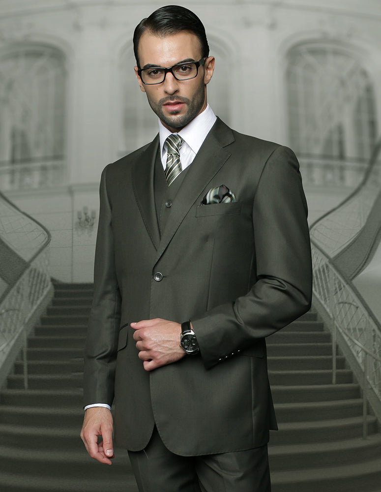 An individual wearing the STATEMENT CLOTHING | TZ-100BG-OLIVE ensemble by Statement Clothing, featuring a dark suit with pleated pants and a tie, stands in front of a grand staircase. He accessorizes with glasses and a watch.