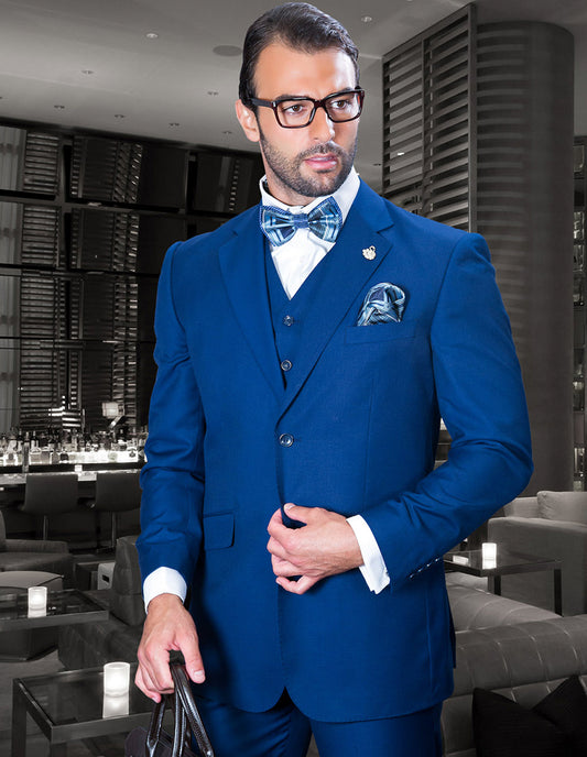 A man in a Statement Clothing TZ-100BG-Sapphire suit, wearing glasses and holding a bag, stands in a modern lounge with leather seating and dim lighting.