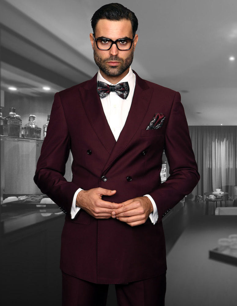 A man wearing the STATEMENT CLOTHING | TZD-100-BURGUNDY from Statement Clothing, complete with pleated pants and a bow tie, stands in a dimly lit room.