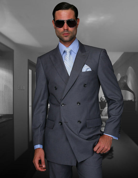 A man in the STATEMENT CLOTHING | TZD-100-CHARCOAL, a tailored gray double-breasted suit by Statement Clothing featuring Super 150's fabric, is paired with a blue shirt and tie. He dons sunglasses and pleated pants while standing confidently in a modern, dimly lit room.