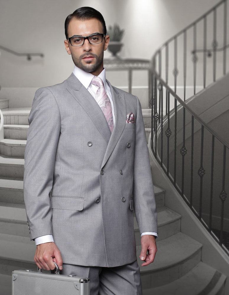 A man in a STATEMENT CLOTHING | TZD-100-GREY from Statement Clothing, complemented by glasses and a pink tie, stands in front of a staircase holding a briefcase.