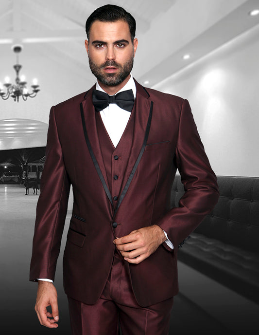 A man in the VENETIAN-BURGUNDY tuxedo by Statement Clothing, featuring black lapels and a bow tie, stands elegantly in a stylish room adorned with chandeliers and plush sofas.